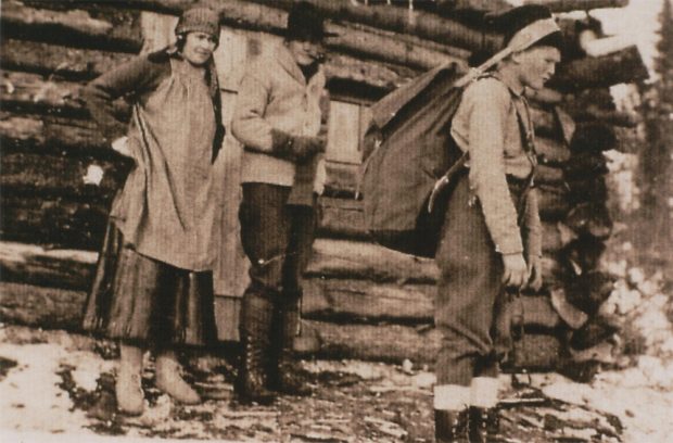 Photo sépia, une femme, un homme et un enfant devant un camp en bois rond, une fine couche de neige recouvre le sol.  L'enfant se prépare à partir, il porte un gros sac à dos, une ganse en cuir aide pour le portage.