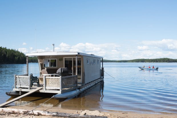 Photo couleur, bateau-maison accosté sur une plage.  Au loin, trois pêcheurs dans une chaloupe.