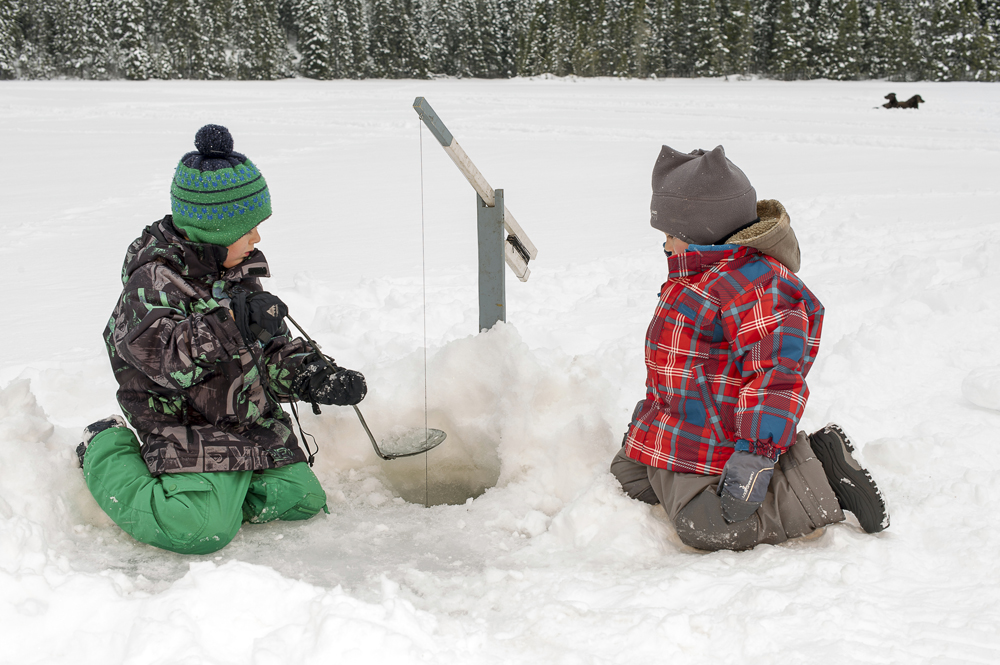 Colour photo, two children kneeling on a frozen lake. One of the children has a ladle to keep the hole from freezing. A strand is placed between the two children.