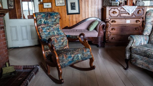 Photo couleur d'une chaise berçante près d'un foyer dans une pièce au décor rustique.  Les accoudoirs se terminent en forme de cygne. Elle est garnie d’un inspirant tissu à motif de chasse.
