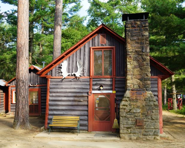 Colour photo, a brown and red log cottage with an immense chimney to the right of the front door.