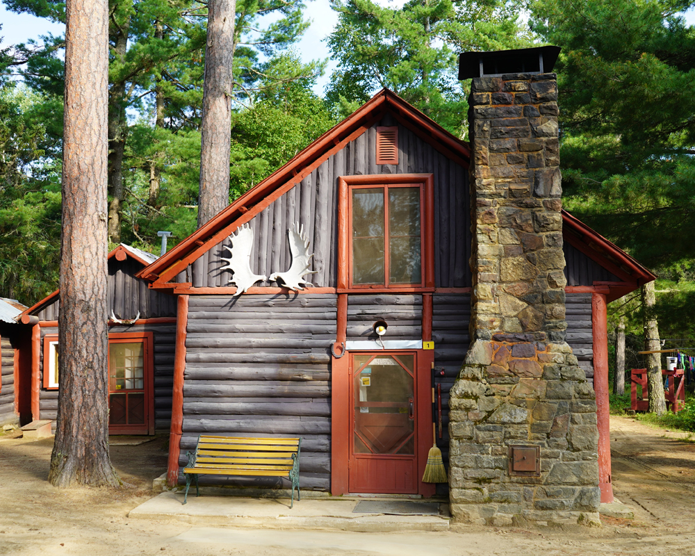 Photo couleur, d'un camp en bois rond brun et rouge ayant une immense cheminée à droite de la porte d'entrée.