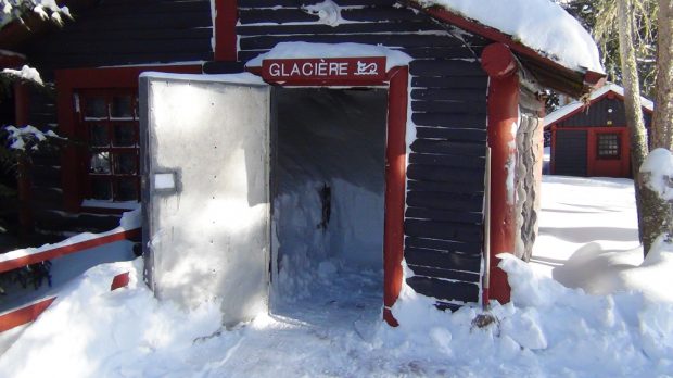 Photo couleur, une petite cabane en bois rond ayant la porte ouverte.  À l'intérieur de la neige entassée.
