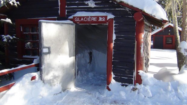 Colour photo, a small log cabin with its door ajar. Inside is packed snow.