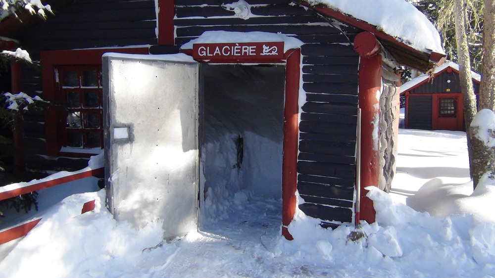 Photo couleur, une petite cabane en bois rond ayant la porte ouverte.  À l'intérieur de la neige entassée.