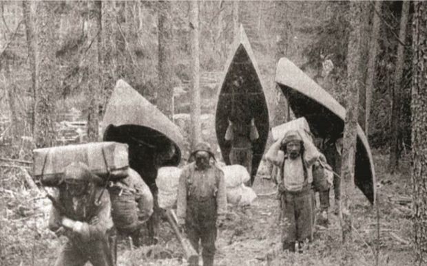 Black and white photo, several men carry canoes and baggage during a portage.
