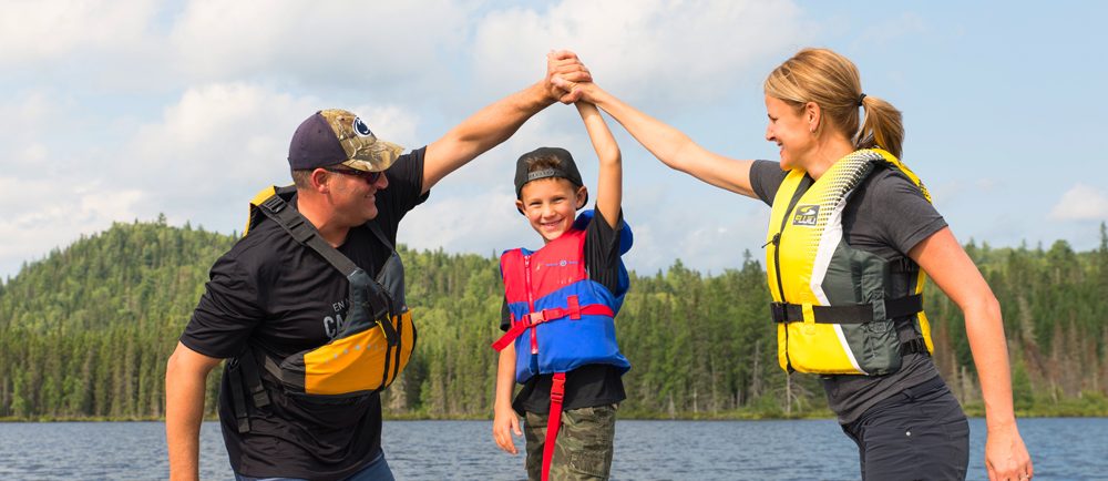 Colour photo, a family of three standing in a rowboat, holding hands and smiling.