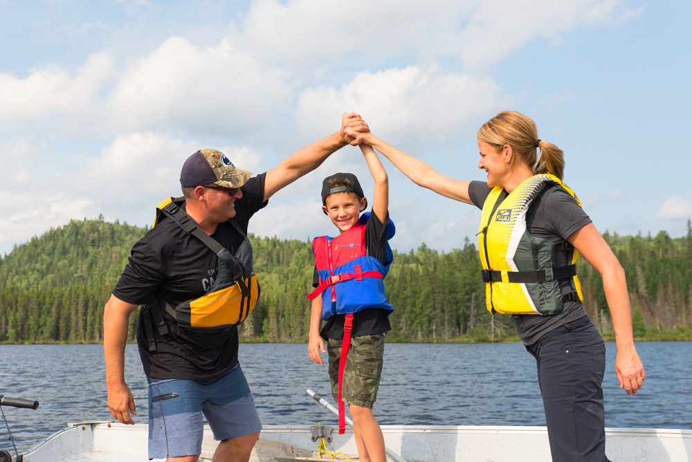 Colour photo, a family of three standing in a rowboat, holding hands and smiling.