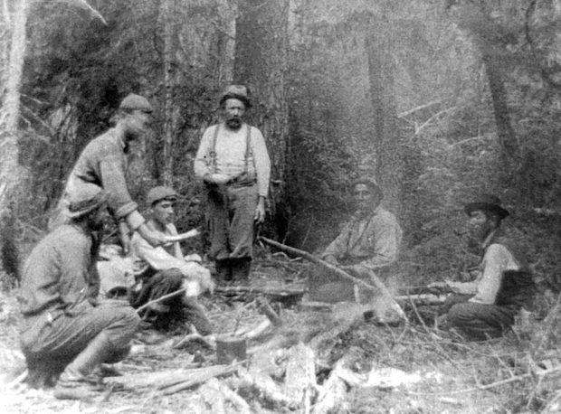 Black and white photo, six men gathered around a fire. Plates in hand, they are getting ready to enjoy a delicious meal prepared by their dedicated guides.