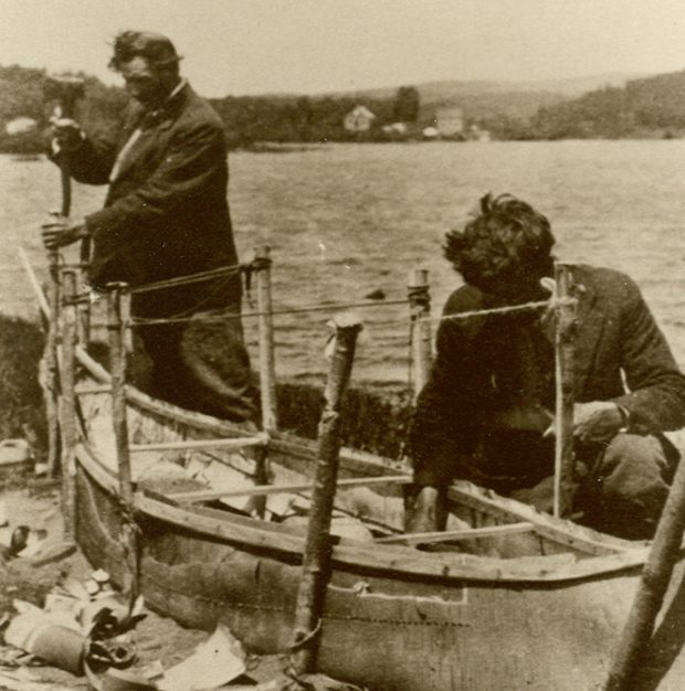 Sepia-toned photo, two men building a traditional birch bark canoe by the shoreline. A few pieces of bark cover the ground.