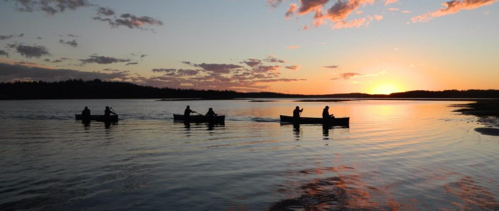 Six paddlers in three canoes glide over the water as the sun sets behind them and the clouds turn pink.