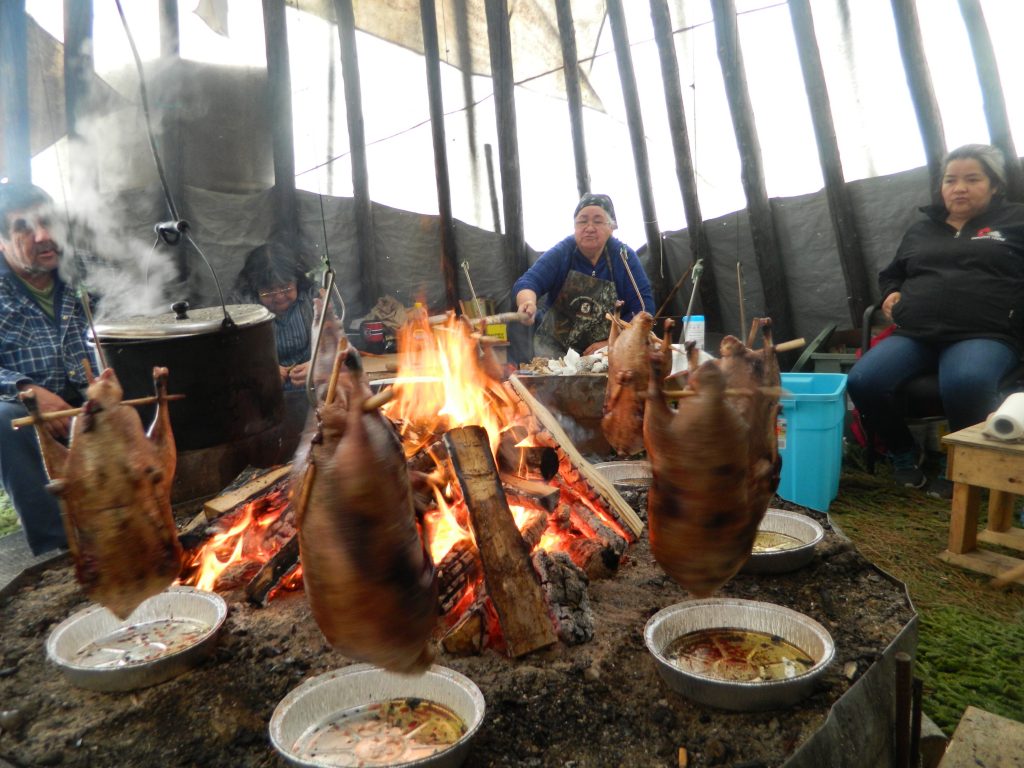 Three people sit behind the fire in a teepee. In the foreground, geese are suspended on cords for roasting.