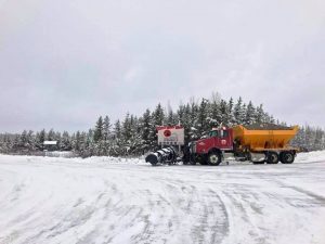 Camion rouge et jaune avec charrue et gravier sur route enneigée.