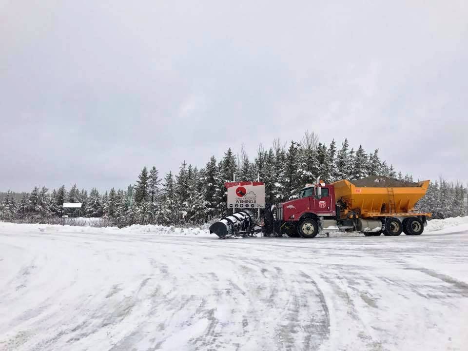 Red and yellow truck with plow and gravel on snowy road.