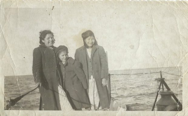 Three girls stand together at the back of a boat.