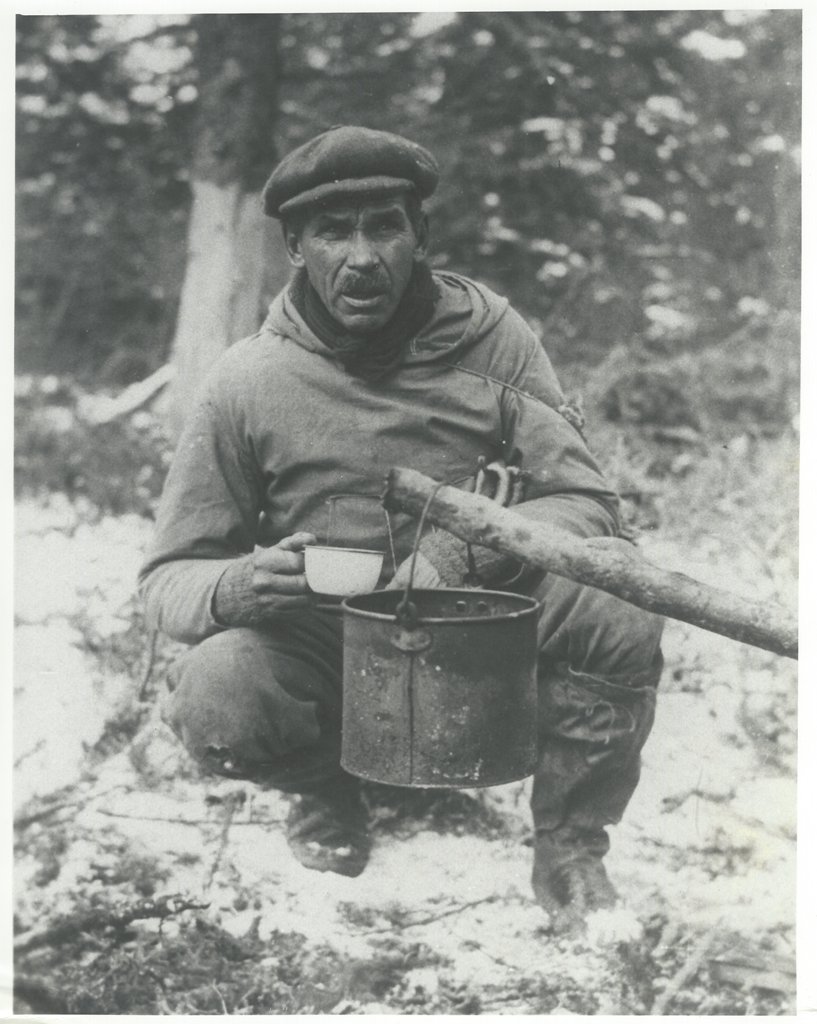 Man with hat and tin cup sitting near forest. He has hung a pail over a fire to boil water.