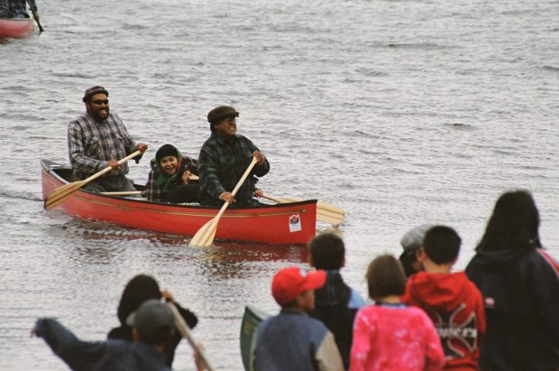 Three people paddling a boat towards children on shore