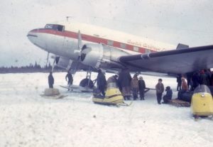 Prop plane on the ice surrounded by skidoos, sleds, and a group of men and children.