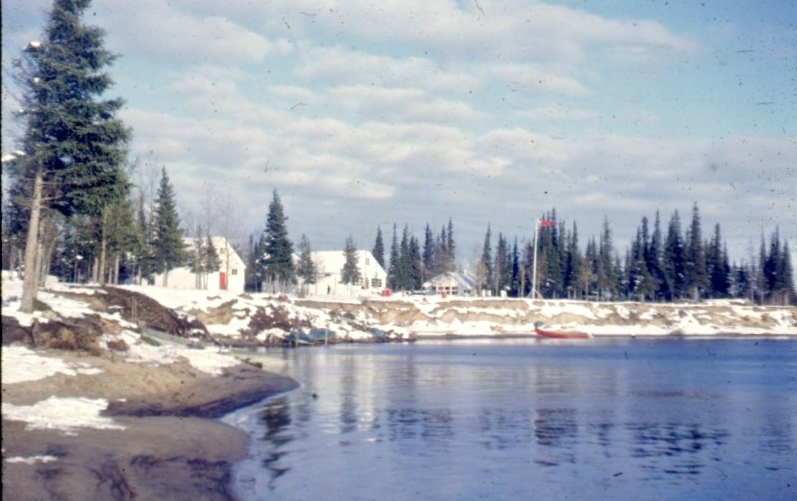 Three white wooden buildings amongst spruces on snowy riverbank.