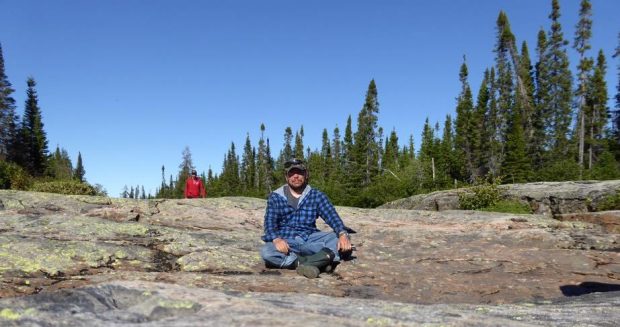 Man in blue checked flannel shirt and jeans sitting in sun on a big rock