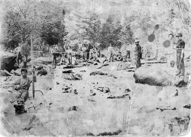 Old black and white photograph of a dozen lumberjacks in the forest. On the left, a man is sitting on a rock and leaning on a peavey.