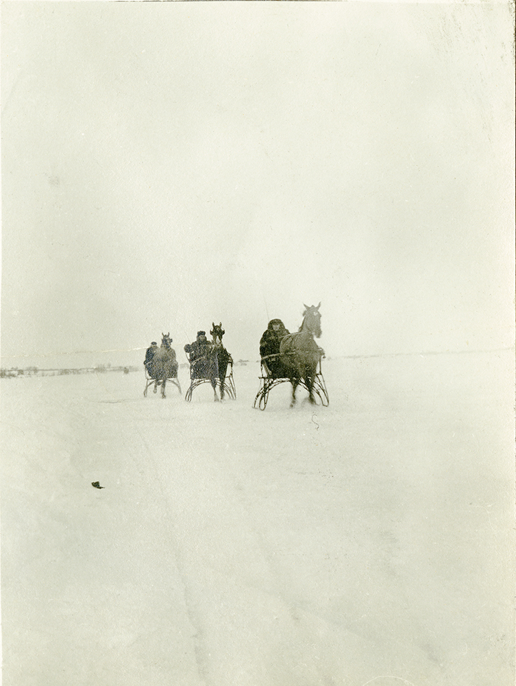 Photographie d’époque en noir et blanc. Trois personnes se déplacent en calèche d’hiver sur une rivière enneigée, solidifiée par la glace.