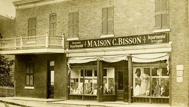 Vintage sepia photograph of a department store display window on the ground floor of a building. The sign says Maison C. Bisson.