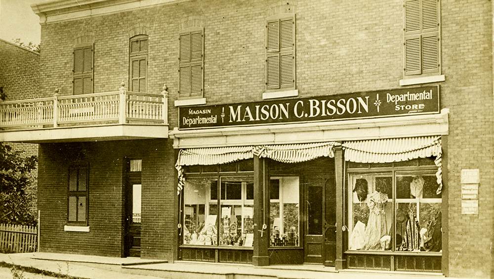 Photographie d’époque en teinte sépia présentant la devanture d’un magasin départemental situé au rez-de-chaussée d’un bâtiment. Sur l’écriteau, on peut y lire : Maison C. Bisson.