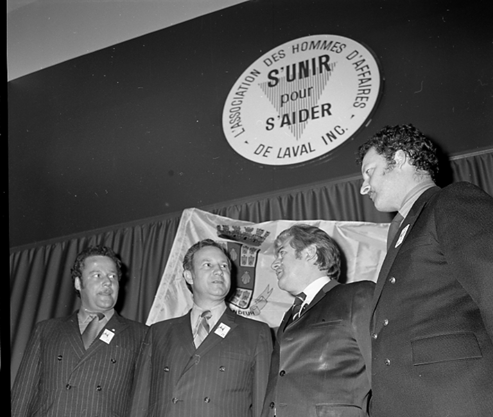 Black and white photograph of four men dressed in black suits talking together. On the wall behind them is a sign that says “L’association des hommes d’affaires de Laval Inc. S’unir pour s’aider” [Laval Businessmen’s Association. Stronger Together].