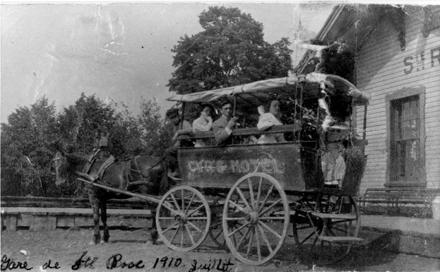 Vintage black and white photograph. It shows two men and three women aboard a calèche in front of a train station. At the bottom of the photograph, the following handwritten words can be seen: Gare de St Rose 1910 juillet [St-Rose station, July 1910].