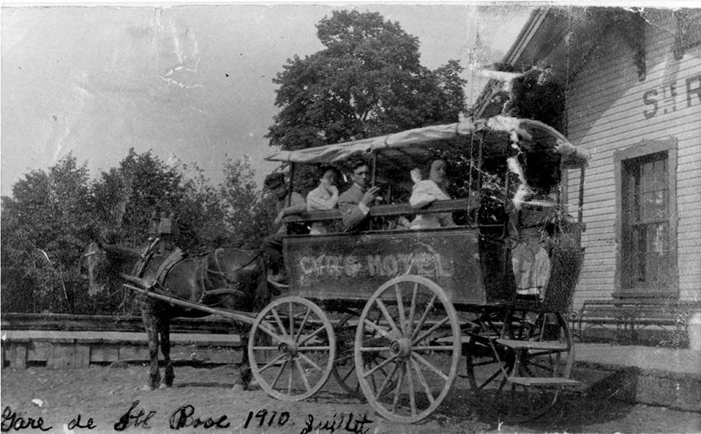 Photographie ancienne en noir et blanc. Elle montre deux hommes et trois femmes à bord d’une calèche, au pied d’une gare. Au bas de l’image, il est écrit à la main : Gare de St-Rose, 1910 juillet. 