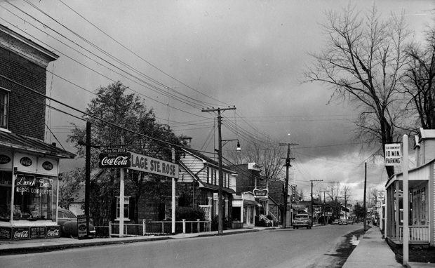 Black and white photograph of a commercial street in the Sainte-Rose district. A few stores, cars parked on the side of the road and the entrance to the Sainte-Rose beach can be seen.