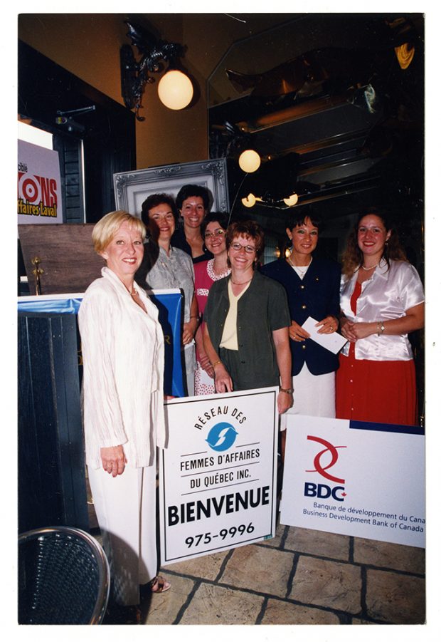 Colour photograph of seven women in a dark room. In the foreground are two signs: one for the Réseau des femmes d’affaires du Québec Inc. and the other for the Business Development Bank of Canada.