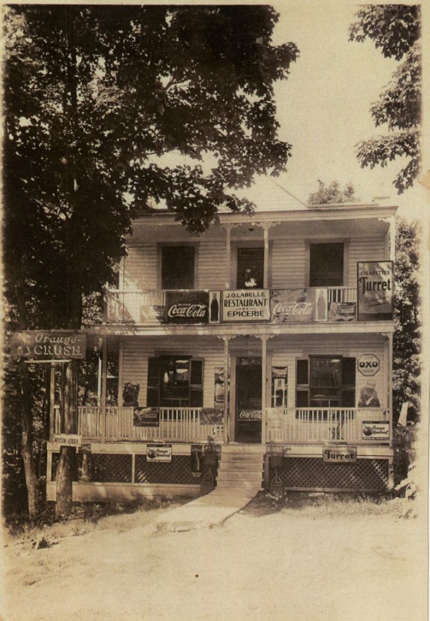 Sepia photograph of a local grocery store. The duplex’s sign reads “J.O. Labelle Restaurant Épicerie.” In front, there are some signs advertising various products, including Coca-Cola and Orange Crush, and Turret cigarettes.