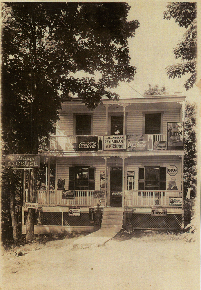 Photographie en teinte sépia d’une épicerie locale. Sur l’écriteau du duplex, il est écrit : J.O. Labelle Restaurant Épicerie. Sur la façade, on peut voir quelques panneaux mettant certains produits, dont les boissons Coca-Cola et Orange-Crush, et les cigarettes Turret.