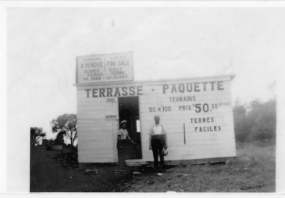 Photographie en noir et blanc montrant deux hommes au pied d’une petite cabane blanche. Il s’agit d’un bureau de ventes de terrains. Le prix est de 50$ pour un terrain de 50 par 100. 