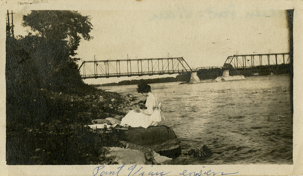 Photographie d’époque en teinte sépia. Elle montre une femme vêtue de blanc assise sur le bord d’une rivière. Au loin, on voit le pont du Canadien Pacifique.