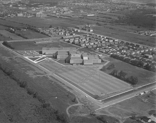 Black and white aerial photograph of Collège Montmorency. The building can be seen in the foreground with a large parking lot surrounded by fields and a small residential neighbourhood.