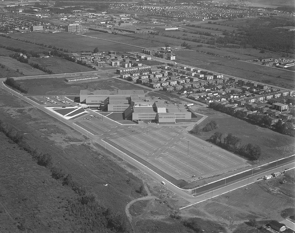 Black and white aerial photograph of Collège Montmorency. The building can be seen in the foreground with a large parking lot surrounded by fields and a small residential neighbourhood.