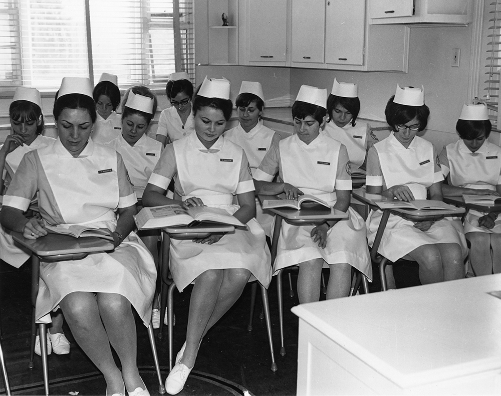 Black and white photograph of 11 women dressed in nurse’s uniforms. They are seated at desks in a classroom, reading textbooks.