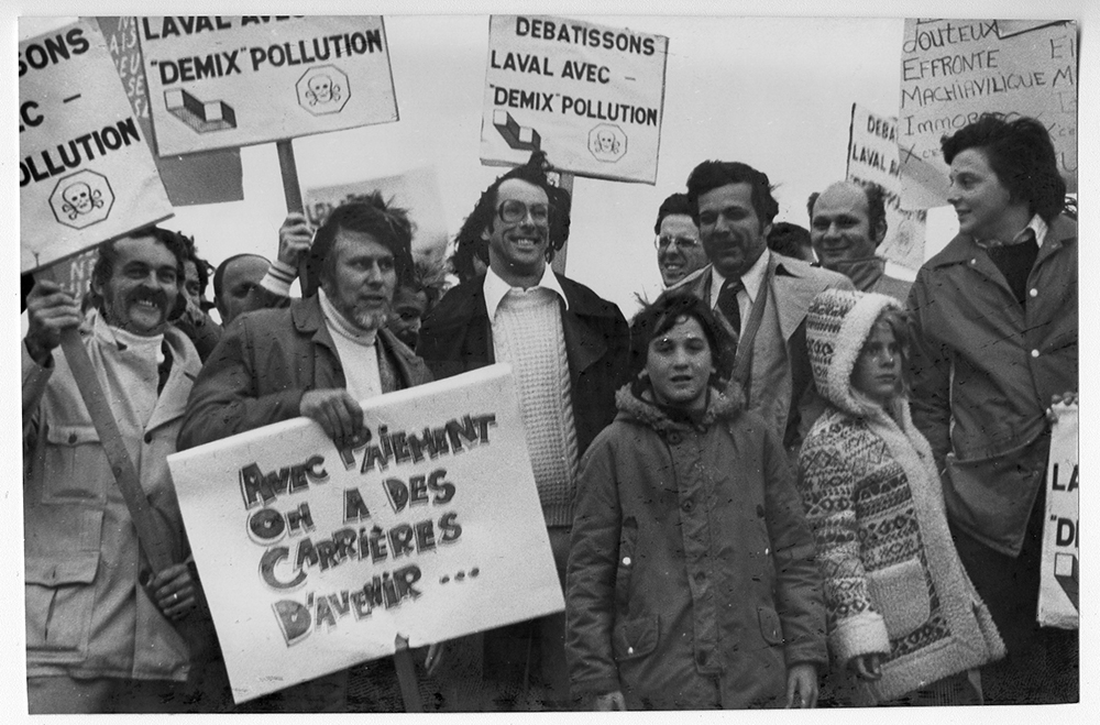 Black and white photograph of citizens demonstrating against the pollution produced by the Demix quarry. In the foreground, a sign held by a man reads “Avec Paiement, on a des carrières de l’avenir” [With (then mayor of Laval) Paiement we have the quarries of the future.]