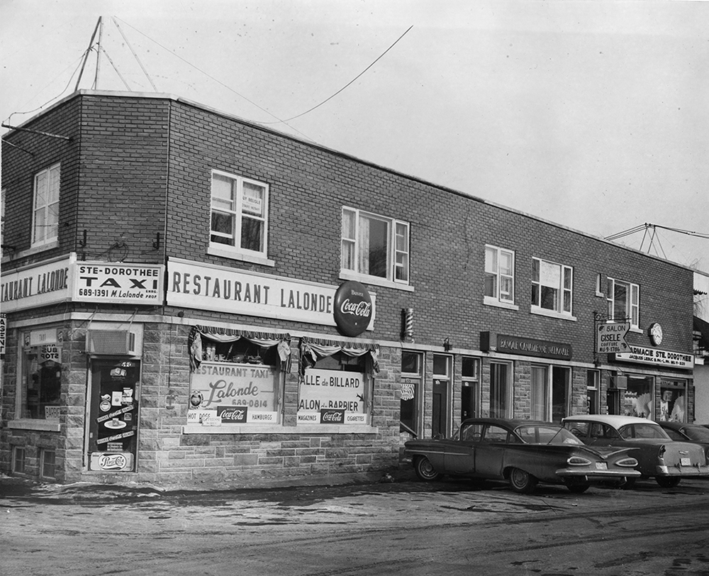 Photographie en noir et blanc d’un bâtiment qui héberge plusieurs commerces : Le restaurant Lalonde, un salon de coiffure et une pharmacie. 