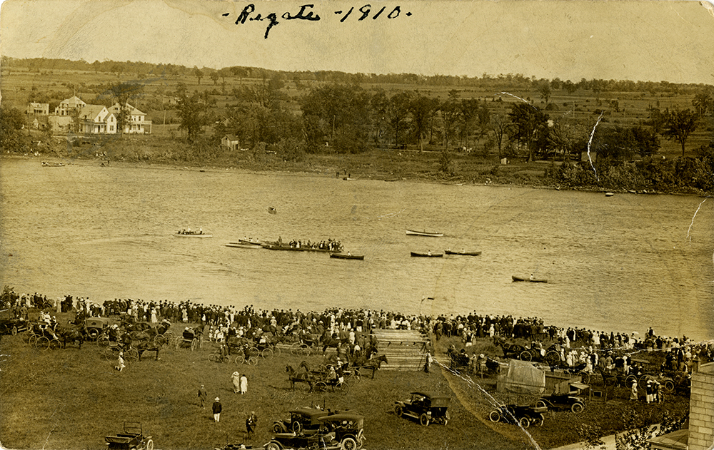 Vintage sepia photograph showing a hundred or so people watching a regatta on a river. At the top of the image are the handwritten words “Régate –1910” [Regatta – 1910].
