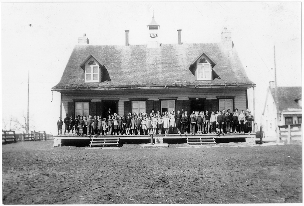 Black and white photograph of a small country schoolhouse. Some 40 students are standing on the front porch.