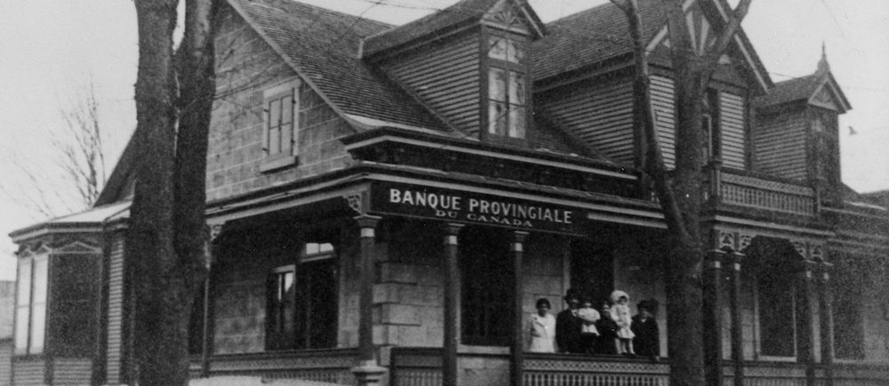 Black and white photo of an ancestral house. It served as a branch of the Provincial Bank of Canada. A family is standing on the front porch.