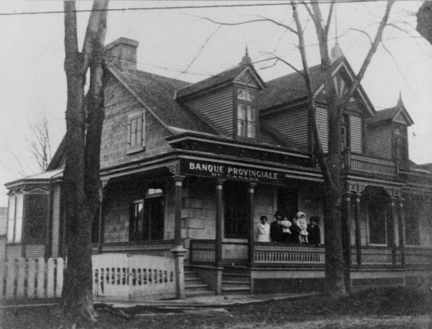 Black and white photo of an ancestral house. It served as a branch of the Provincial Bank of Canada. A family is standing on the front porch.