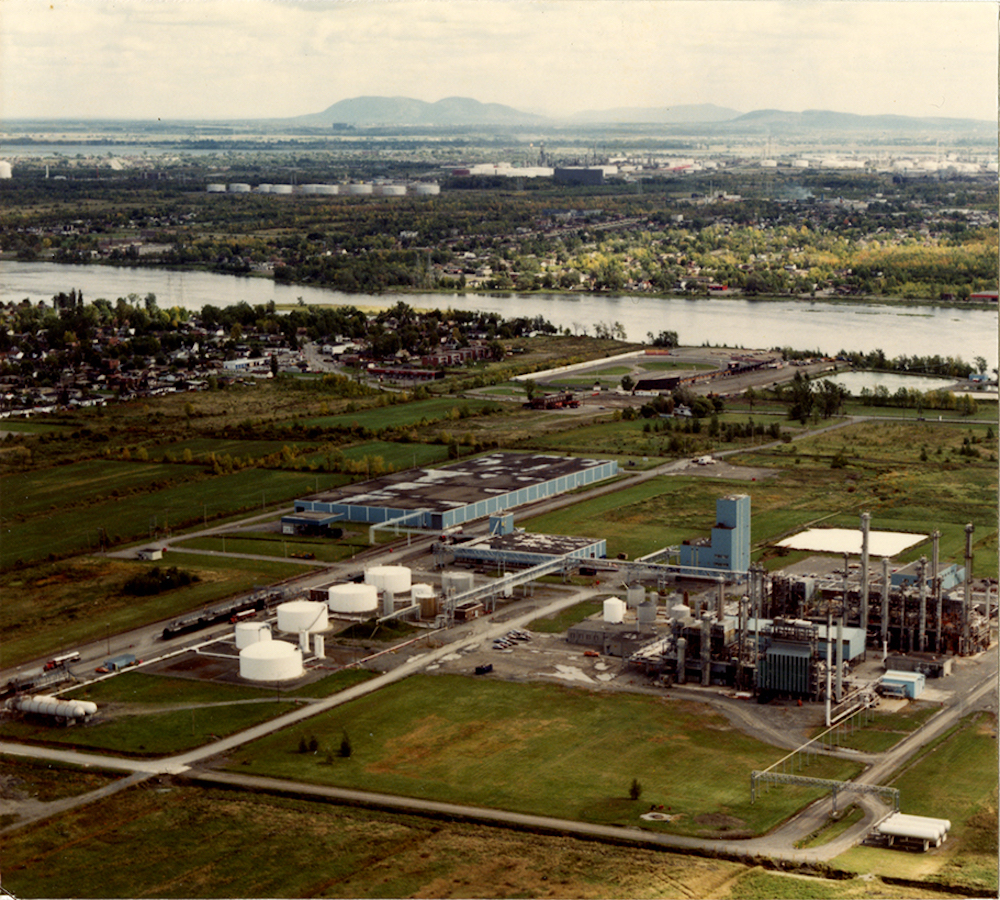 Photographie aérienne en couleur d’une ancienne raffinerie à St-François, installée près d’une rivière et d’un quartier résidentiel. 