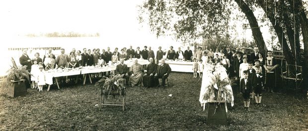 Black and white picture of a large group gathered around a table outside. There is a stretch of water in the back. Bishop Georges-Marie and his brother Armand are in front of the group with 2 other men.