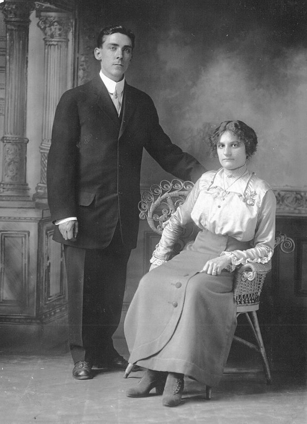 Black and white picture of Emma LePailleur sitting in a rattan armchair and Joseph Belisle standing next to her. Behind them, a painted decoration suggests Greek columns.