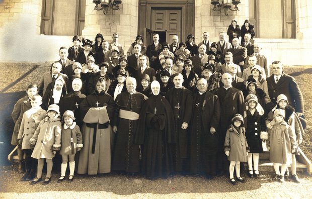 Black and white picture of a group of about 50 people in front of a church. 8 religious are in the foreground, including Bishop Georges-Marie and Sister Agnès.